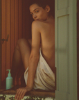 A woman wrapped in a towel sitting in an open window, looking over her shoulder, a CORPUS body cream pump bottle beside them on the sill