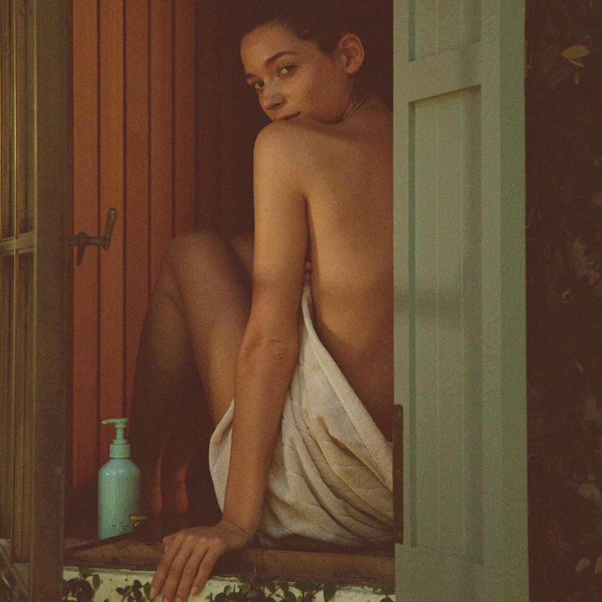 A woman wrapped in a towel sitting in an open window, looking over her shoulder, a CORPUS body cream pump bottle beside them on the sill